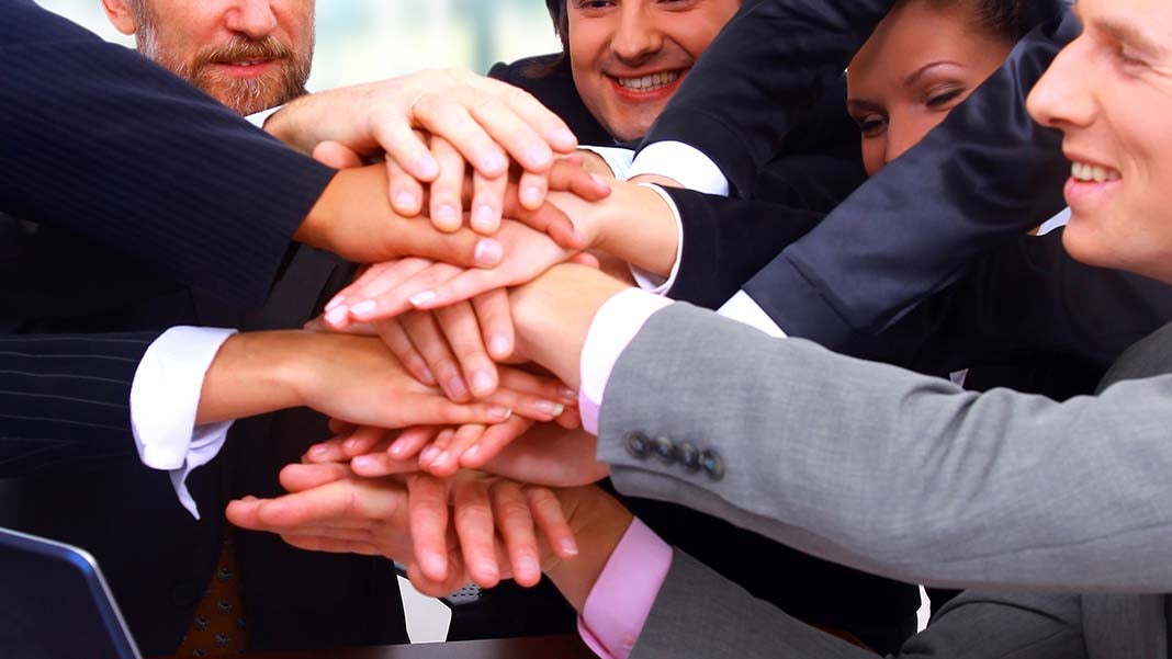  A group of diverse business professionals in suits joining their hands together over a laptop, symbolizing unity, teamwork, and support in a positive work environment.