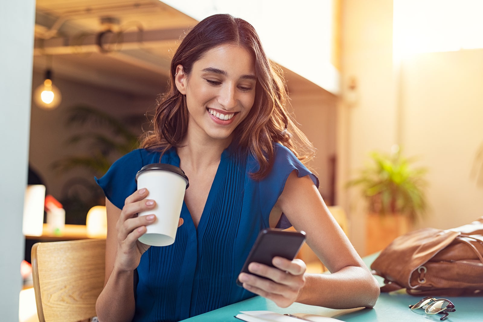 Young beautiful woman holding coffee paper cup and looking at smartphone while sitting at cafeteria. Happy university student girl using mobile phone. Businesswoman drinking coffee and smiling.