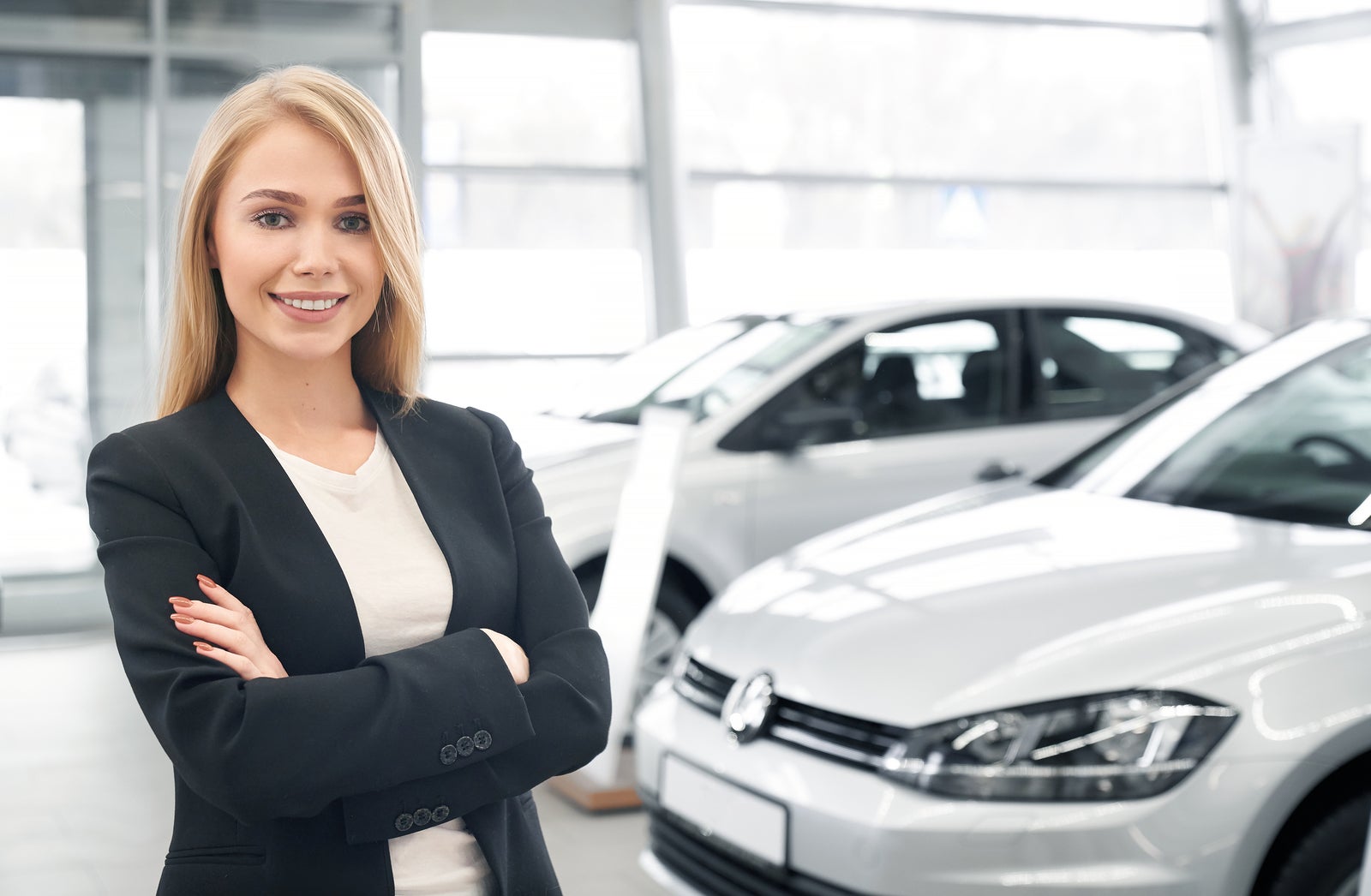 Smiling female car dealer with folded arms looking at camera and posing in front of new automobiles. Pretty woman wearing smart suit working in auto salon and selling cars to clients.