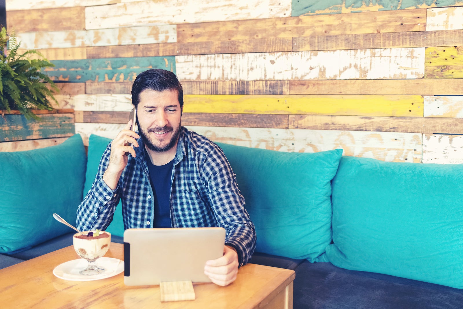 Happy mature bearded entrepreneur doing remote job talking on mobile phone while working on digital tablet in trendy cafe - modern freelancer man surfing net while enjoying dessert.