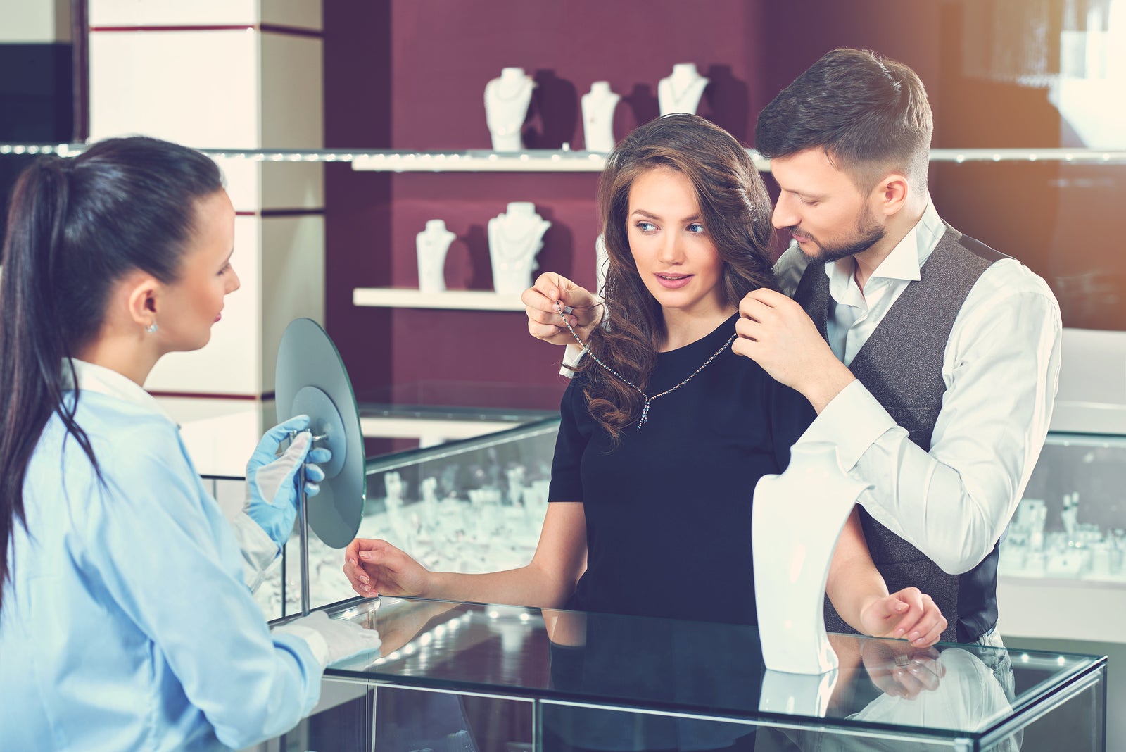 Handsome young man trying necklace for his gorgeous wife at jewelry store. Jeweler in gloves holding mirror for buyer. Brunette woman with wavy hair, wearing black dress estimating looking at mirror.