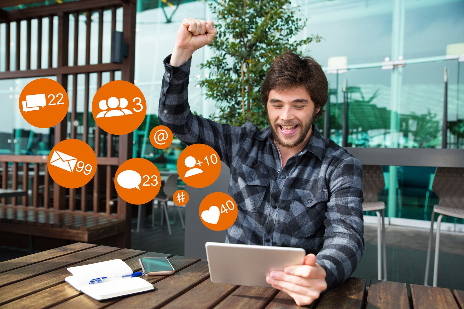 Happy man using tablet and celebrating achievement in cafe with social media icons. Attractive guy raising clenched hand and sitting at wooden table. Success concept. Front view.