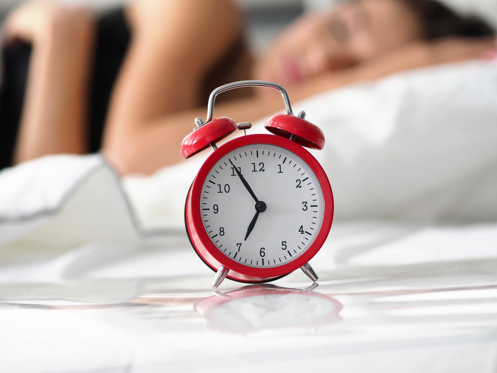 Red old fashioned alarm clock set at seven in morning while woman sleeping at background in her bed closeup