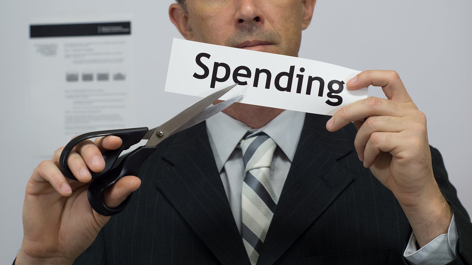 Male office worker or businessman in a suit and tie cuts a piece of paper with the word spending on it as a spending or expense reduction business concept.
