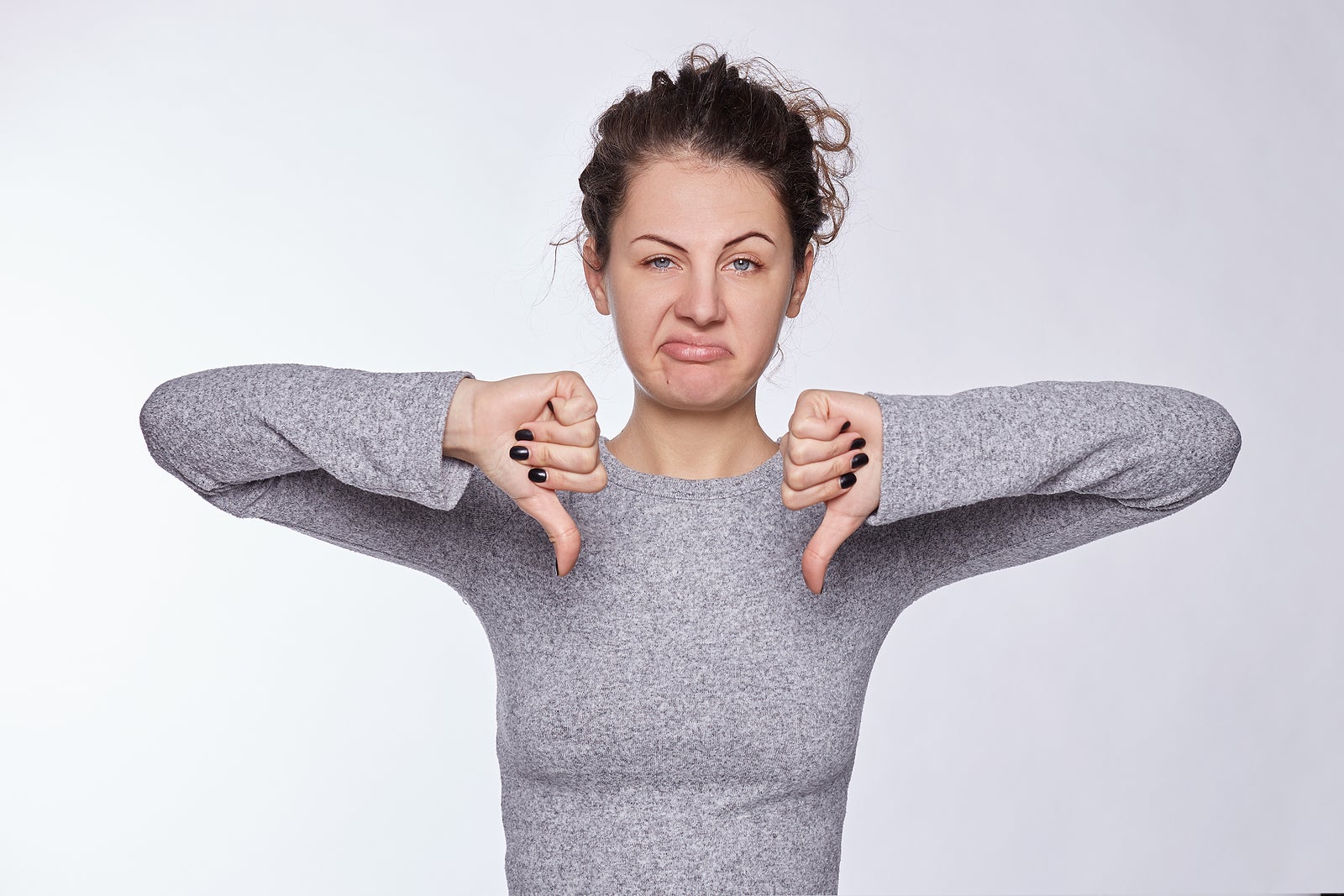 Curly haired Caucasian pretty female expresses dislike with body language, keeps thumbs down and frowns face, has displeased face, curved  lips, isolated over white background. This is really bad idea!