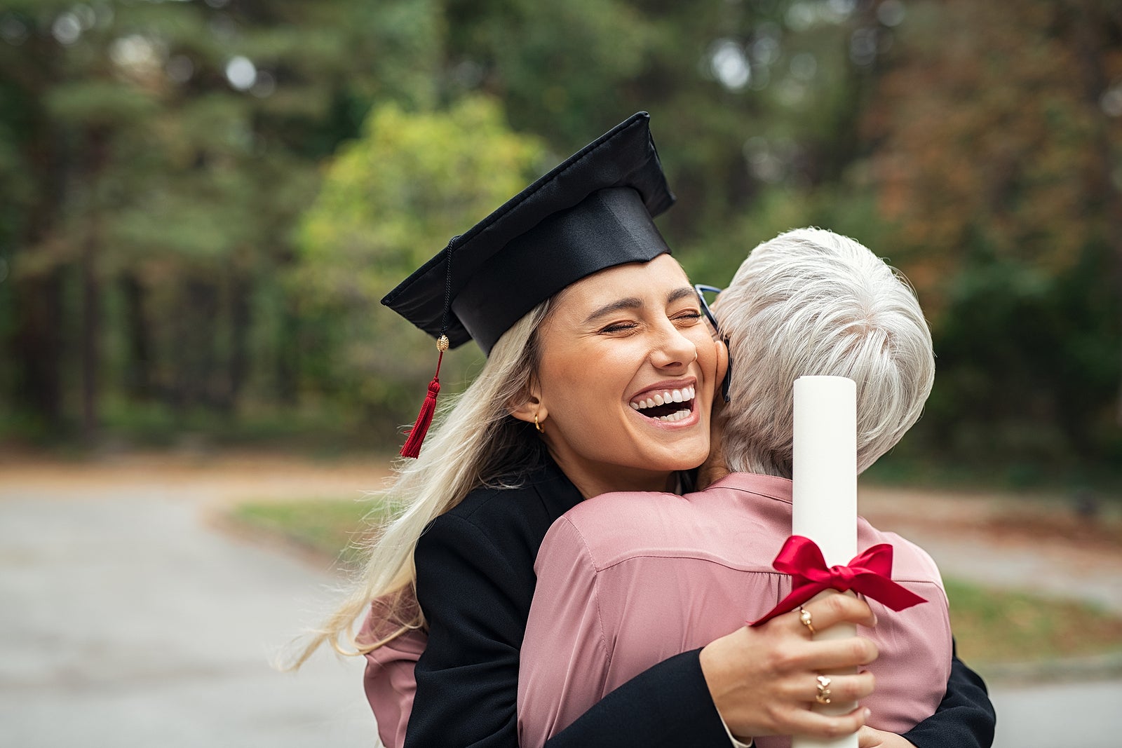 Enthusiastic graduated daughter holding degree hugging mother in campus. Young female student graduate hugging her mom at graduation ceremony. Excited college student with the graduation gown and hat.