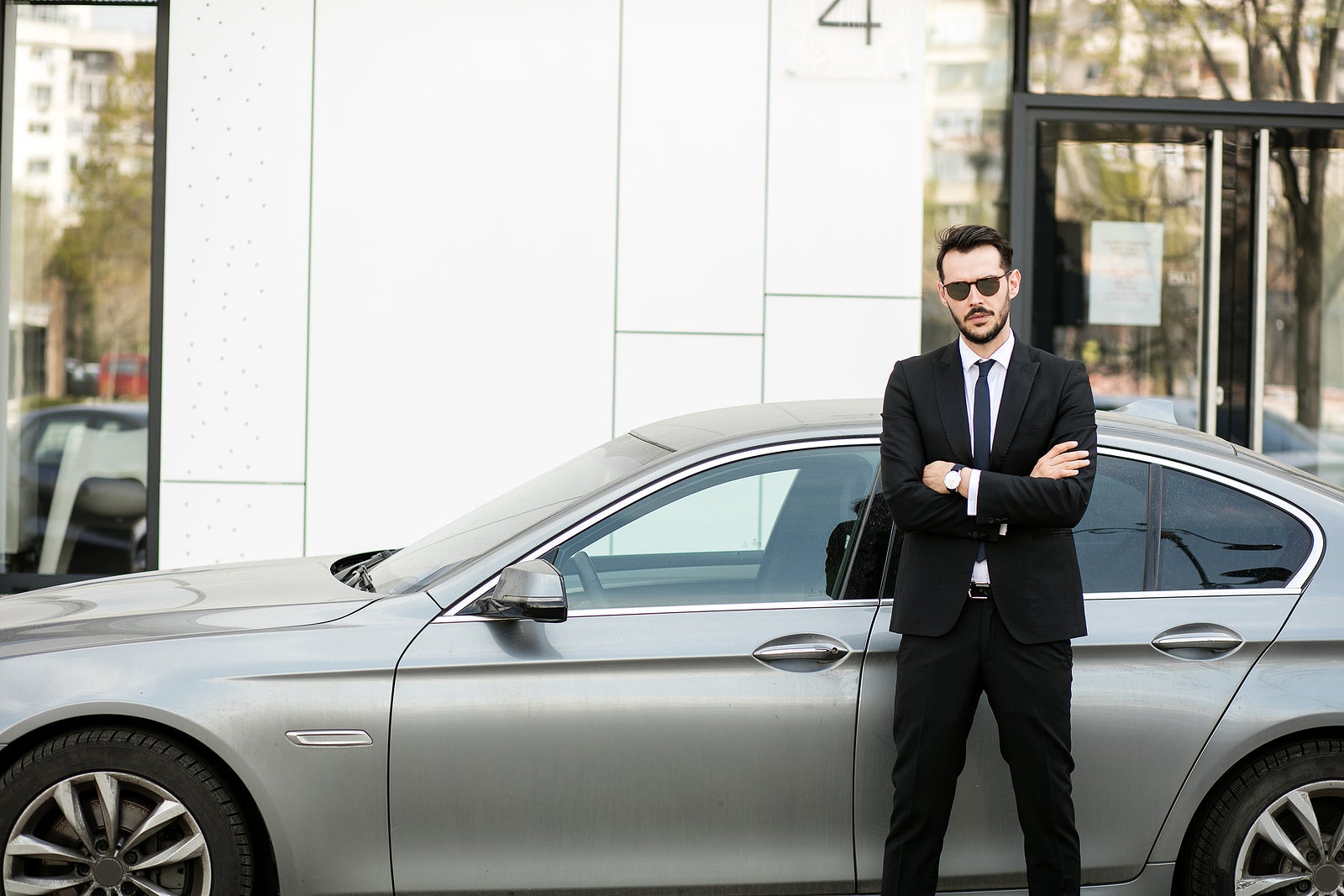 uber driver in elegant suit in front of an luxury car showing his cellphone, outside on the street in front of an office building