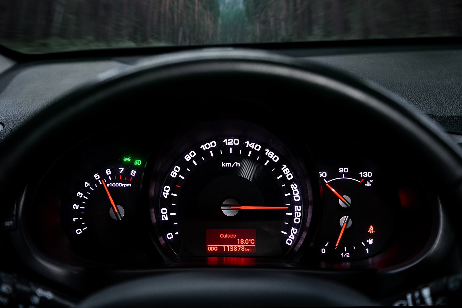A close-up of a car control panel with a speedometer indicating a huge speed of 220 km per hour, a tachometer at 5000 rpm, oil and gasoline levels. The car rides on the highway at high speed along the forest