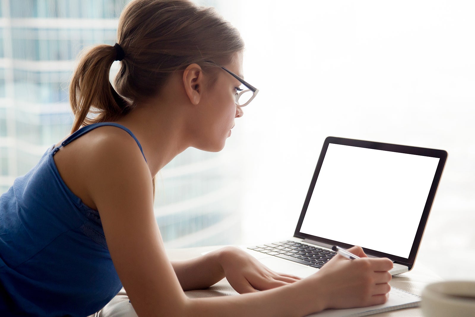 Young serious woman wearing glasses looking at pc laptop white blank mock up screen advertising product or service, writing down some notes, online study, job search, hotel booking, place for ads