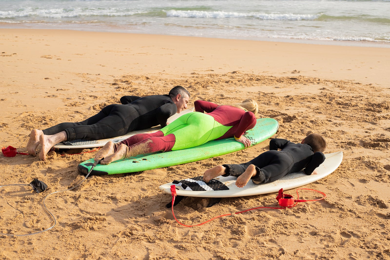 Family of surfers training on sand beach. Mother, father and son in full body swimsuits laying on surfboards. Sea waves on background. Back view. Vacation, surfing and summer concept