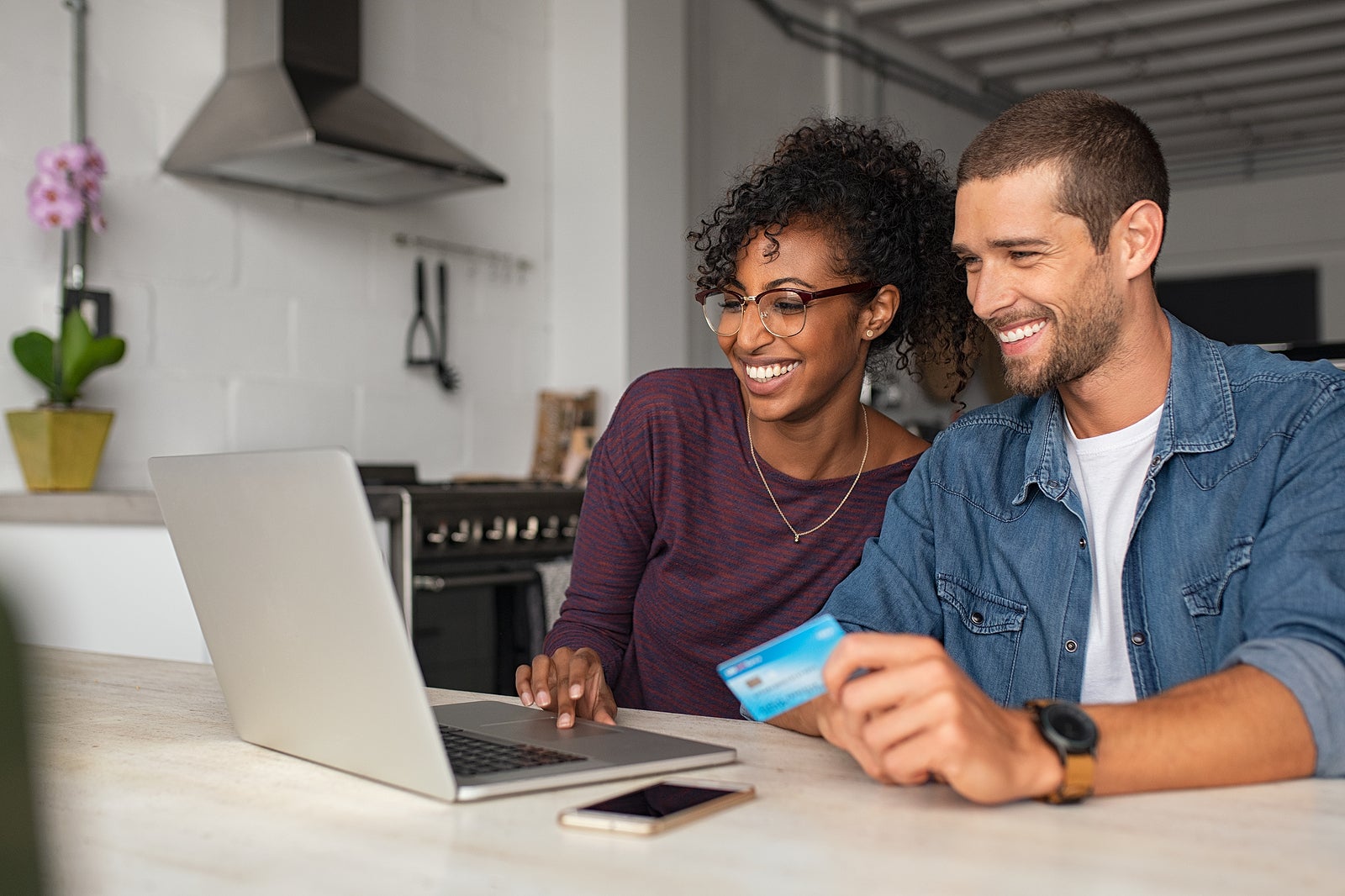 Smiling young couple making shopping online with credit card and laptop at home. Happy multiethnic couple holding debit card while buying on ecommerce site using laptop.