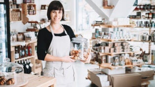 Small local business owner. Seller assistant with glass jar of pastries in interior of zero waste shop. Cheerful woman in apron stands behind counter with food products in plastic free grocery store.