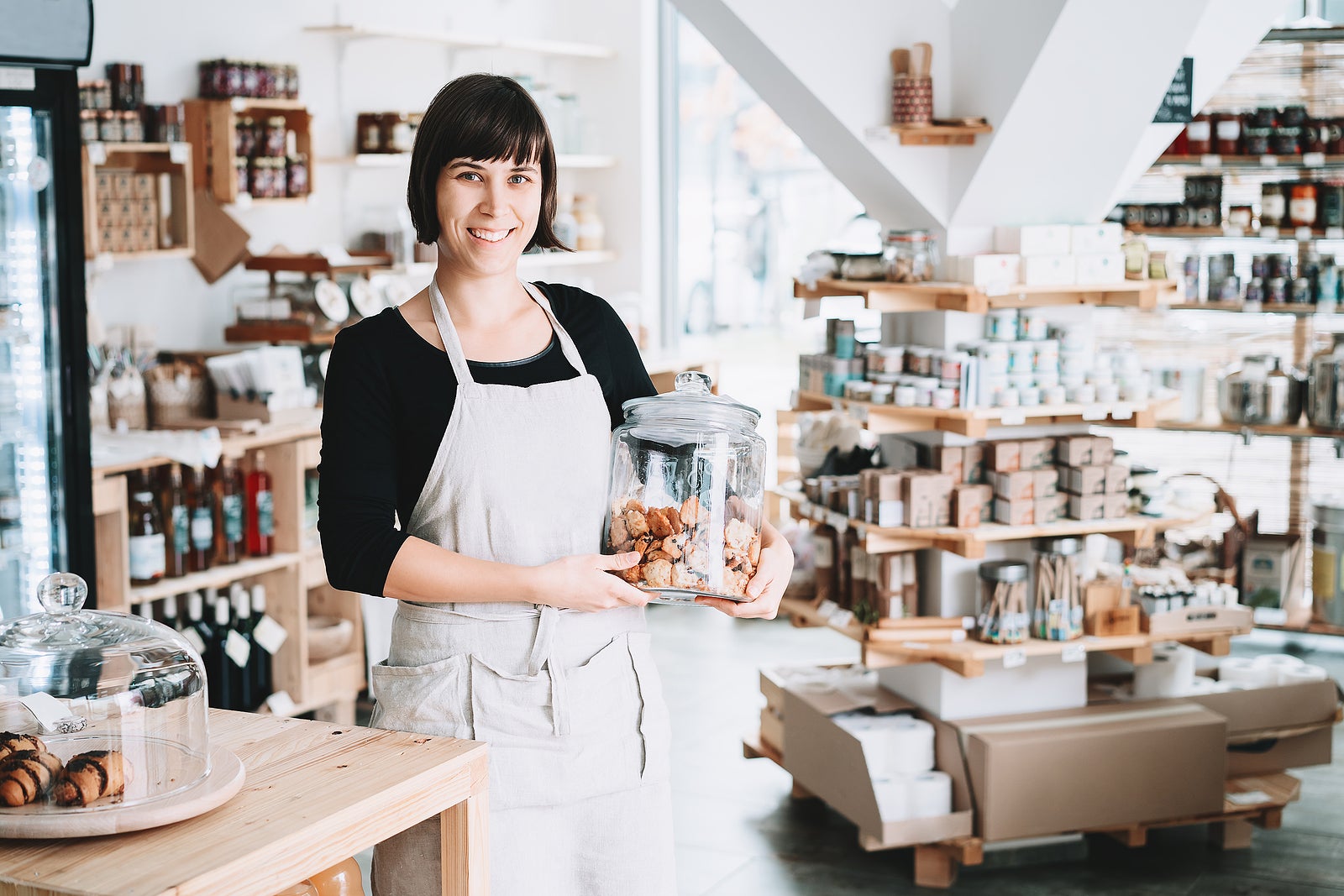 Small local business owner. Seller assistant with glass jar of pastries in interior of zero waste shop. Cheerful woman in apron stands behind counter with food products in plastic free grocery store.