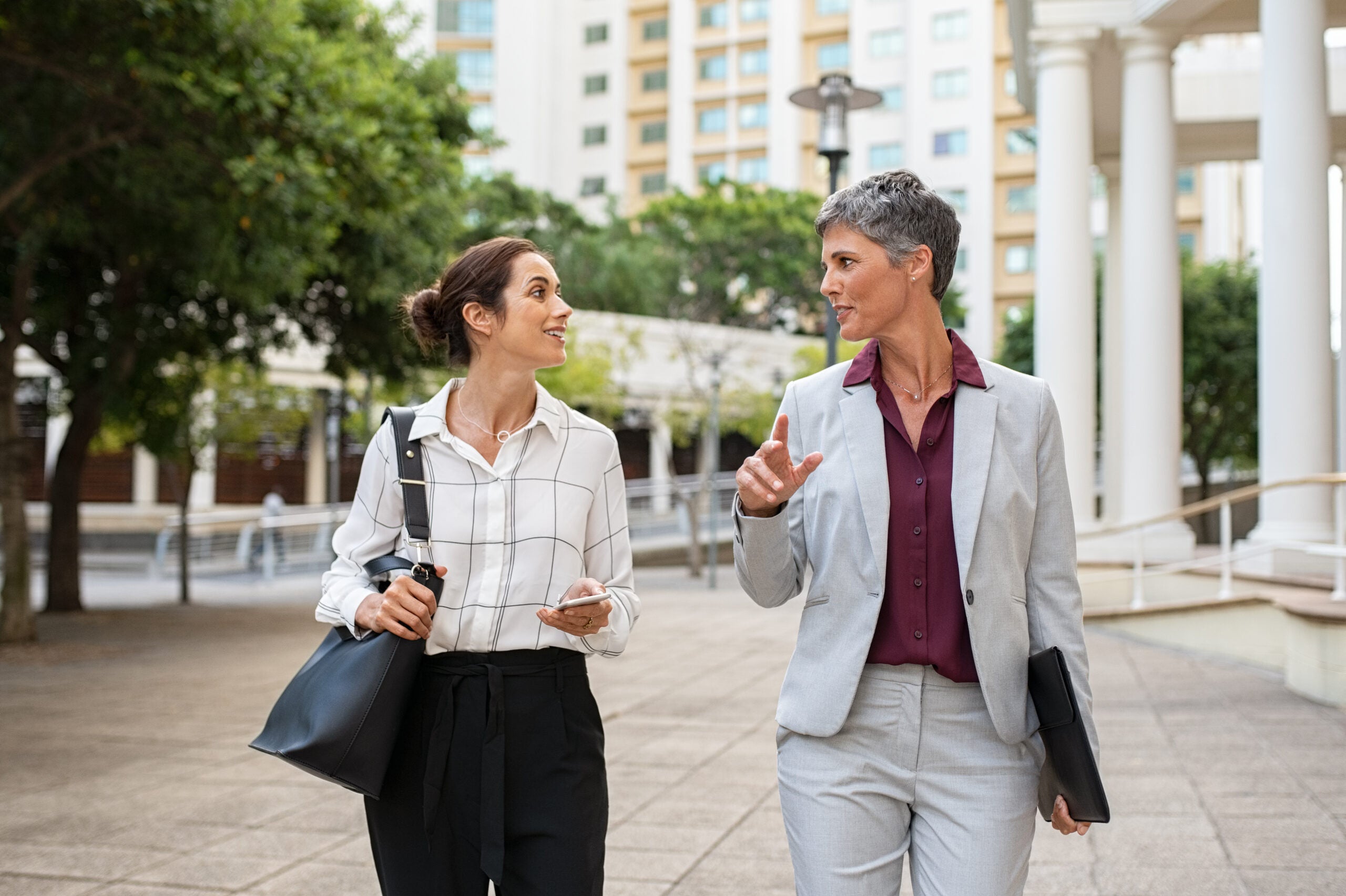 Two business women in conversation walking on city street. Corporate colleagues discussing new project while going to work. Two mature formal women discussing business outdoor.