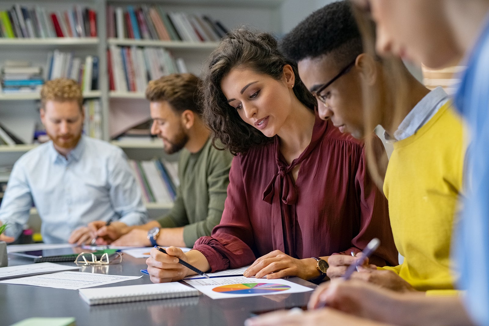 Businesswoman training young business man while explaining graph. African businessman analyzing report with team. Group of multiethnic creative business people sitting while working at financial plan.