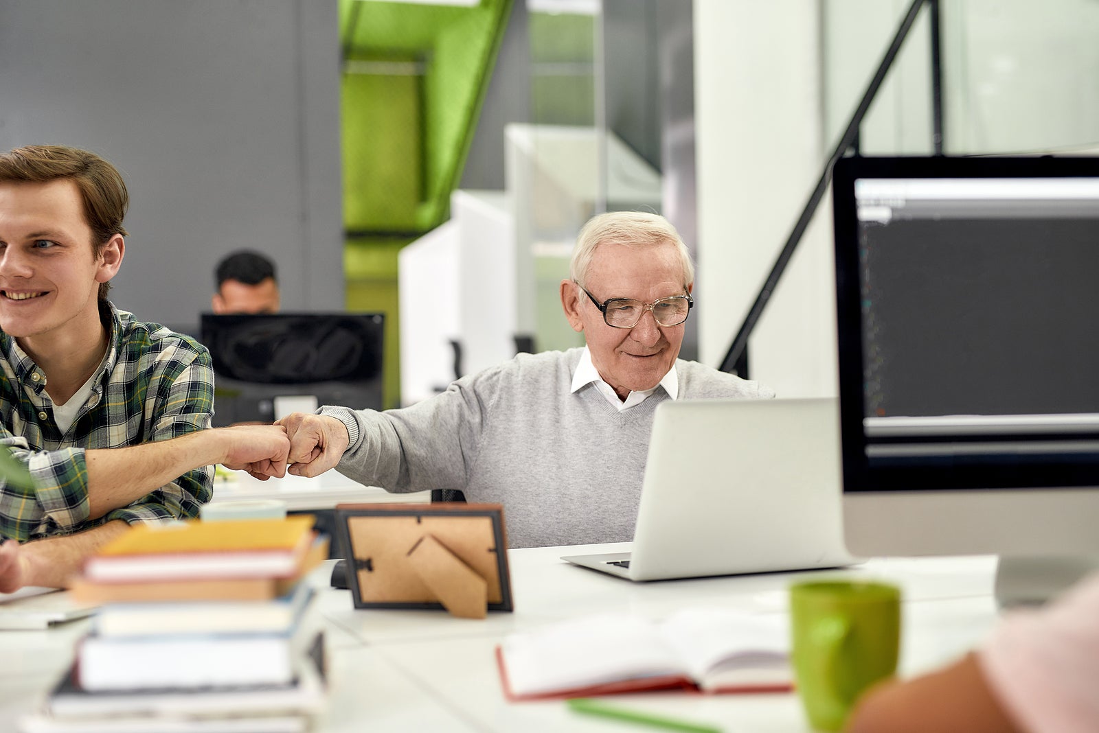 Aged man, senior intern looking at the screen of his laptop and doing a fist bump with colleague, Friendly male worker engaging new employee, working in the office, Selective focus, Horizontal shot