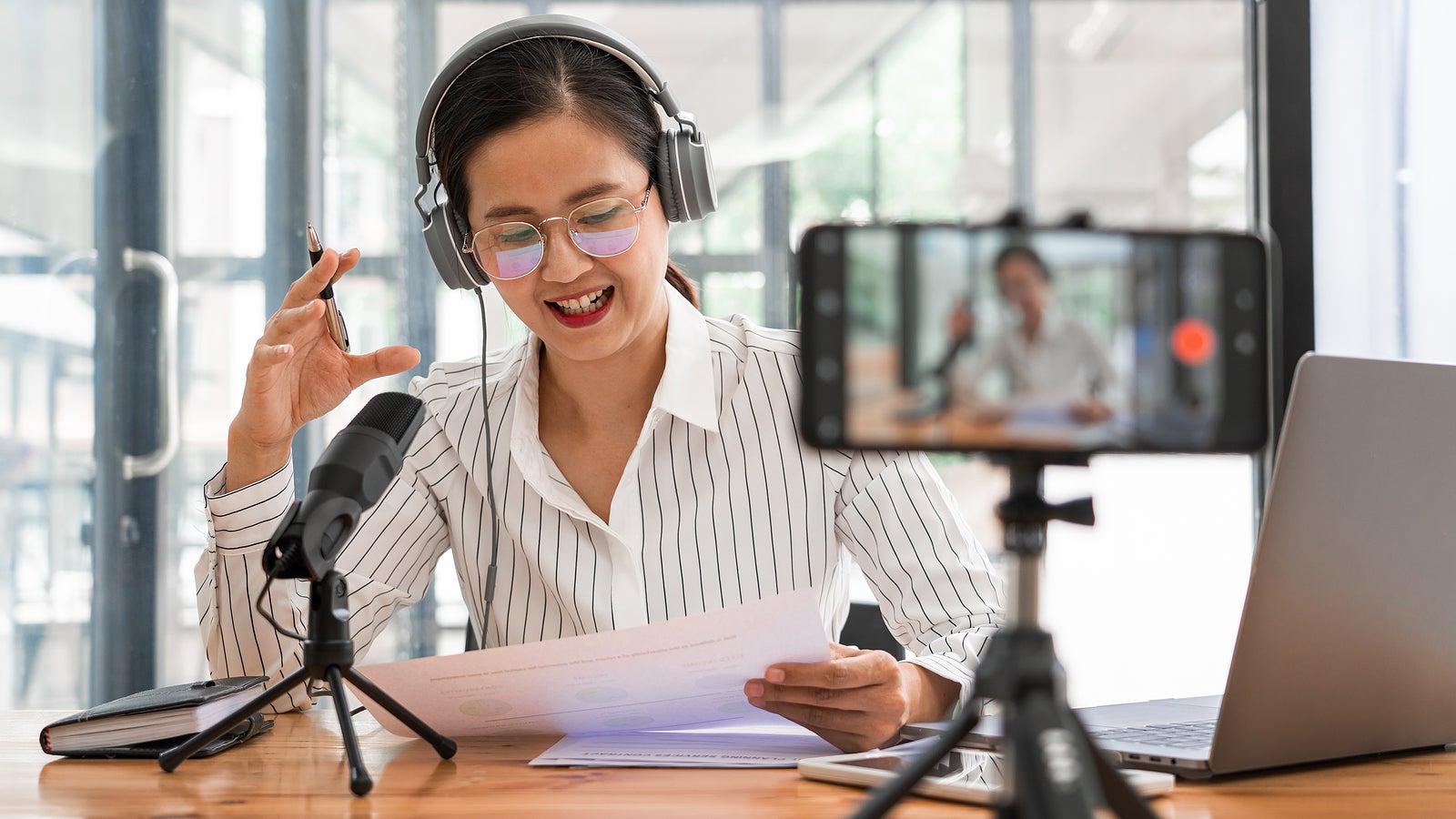 Asian women podcaster podcasting and recording online talk show at studio using headphones, professional microphone and computer laptop on table looking at camera for radio podcast.