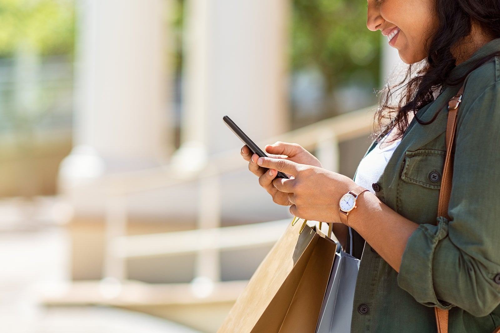 Closeup of woman hands holding shopping bags and using smartphone outdoor. Shopaholic girl checking online with smart phone shopping deals. Young stylish woman using mobile phone for ecommerce.
