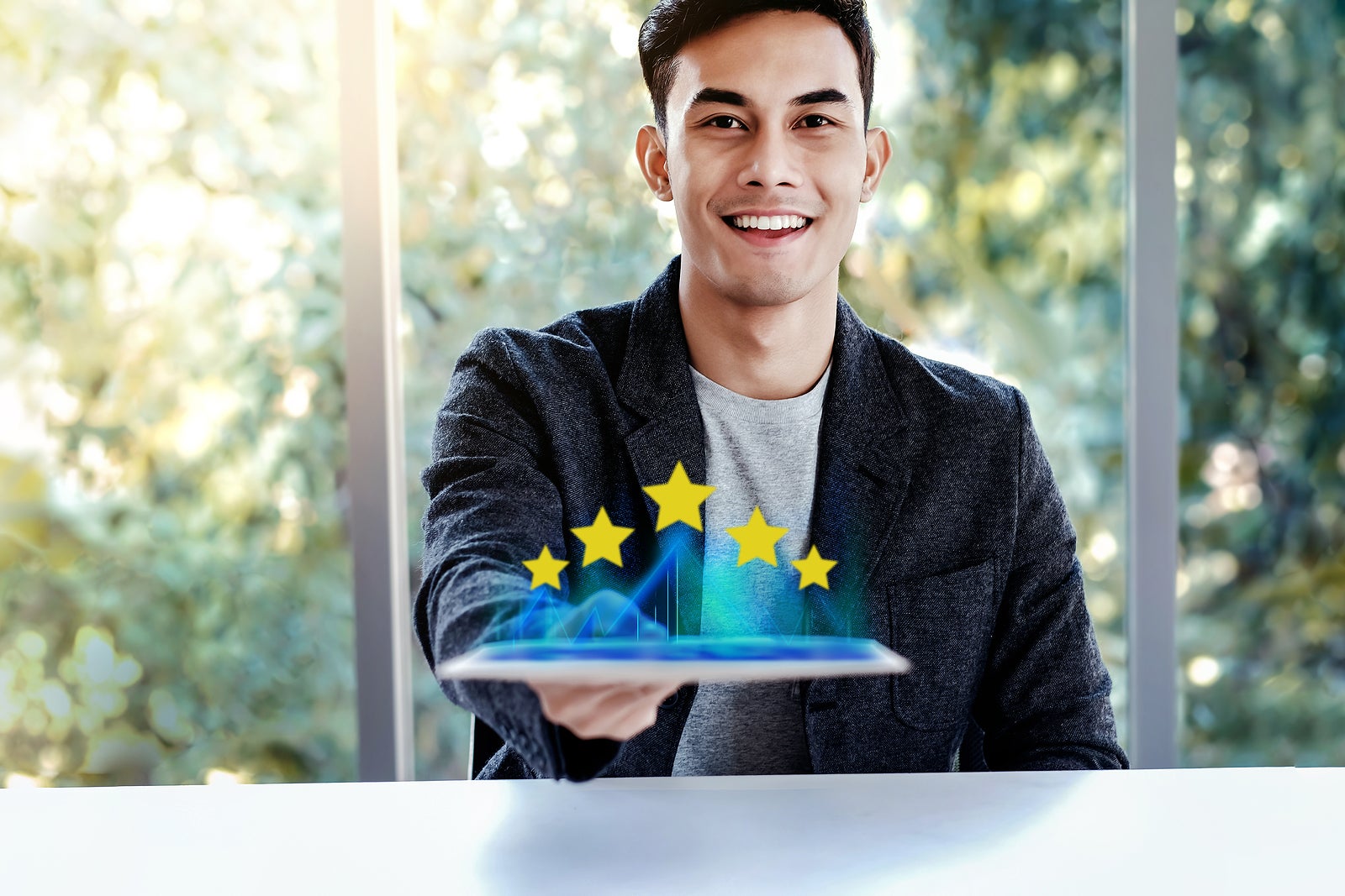 Customer Experiences Concept. Happy Young Man Sitting at the Desk and presenting his Five Star Rating in Online Survey via Digital Tablet. Smiling and Looking at Camera