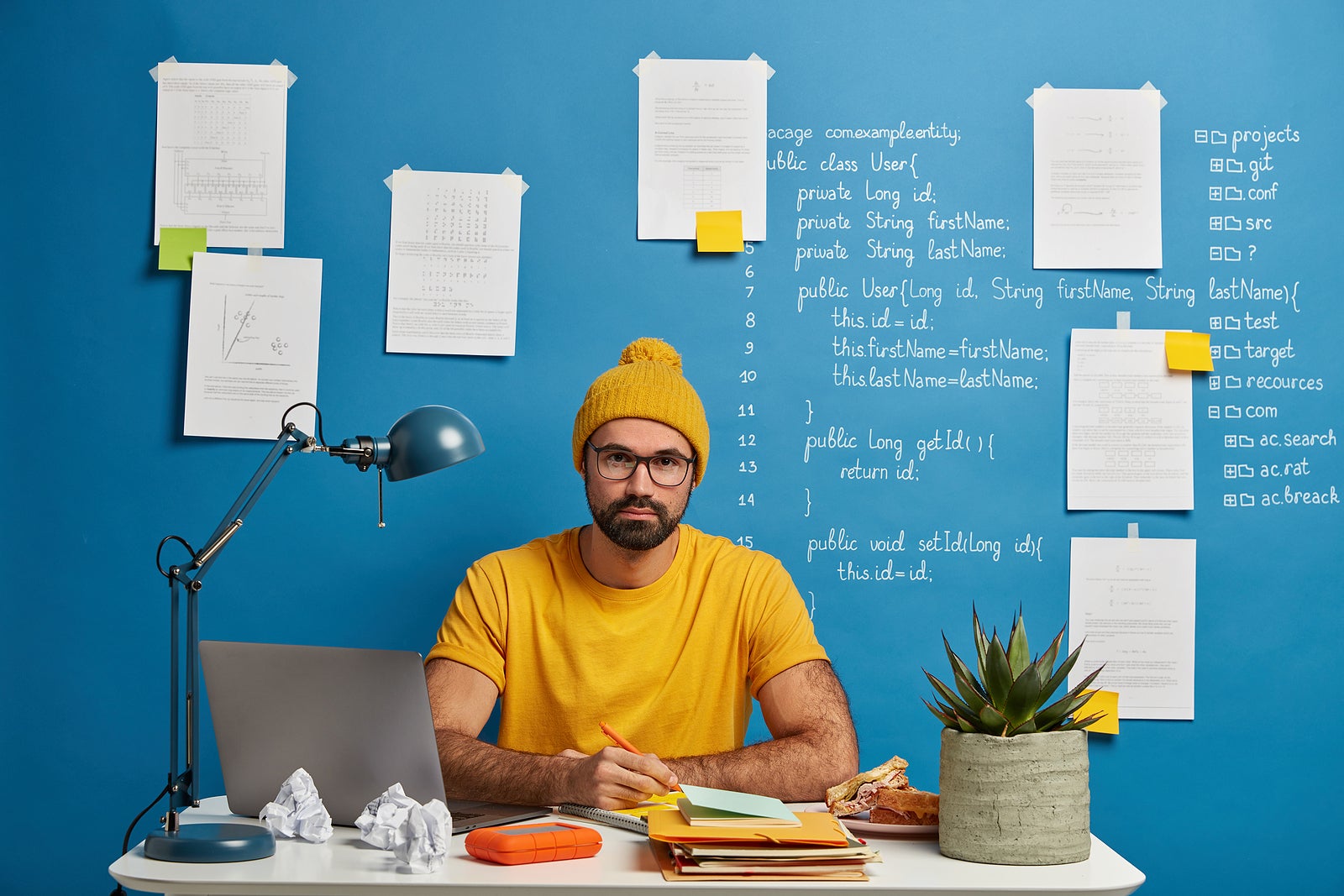 Serious student going to watch training webinar, works on coursework plan, creats article in notepad, wears yellow hat, t shirt and glasses, makes schedule of exam preparation, poses at workplace