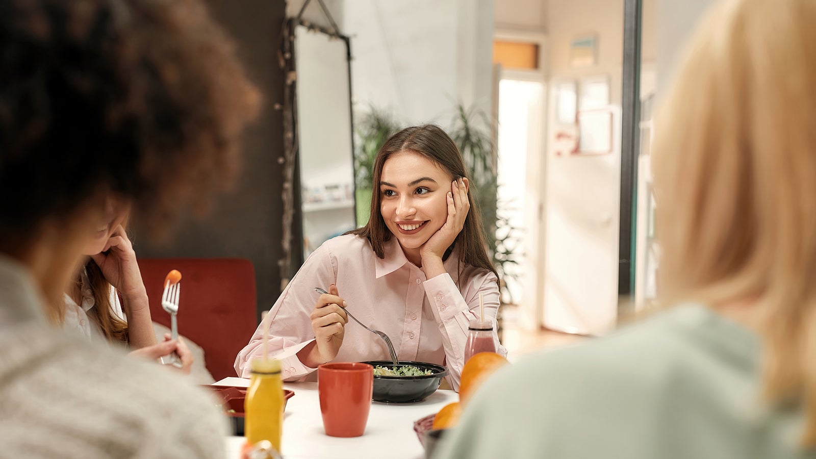 Business lunch. A fashionably dressed young brown haired woman smiling and sitting at a table with her female collaborateurs during a business lunch in a well-lighted creative office