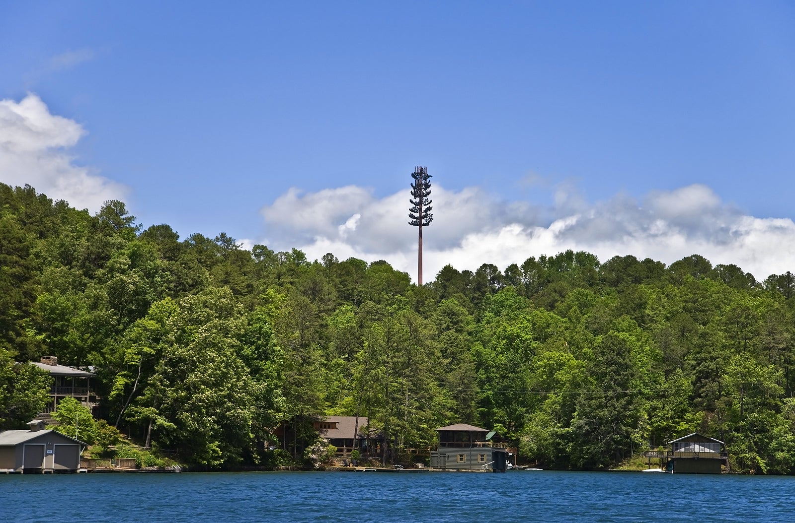 A cell tower high above the tree line over homes and a lake.