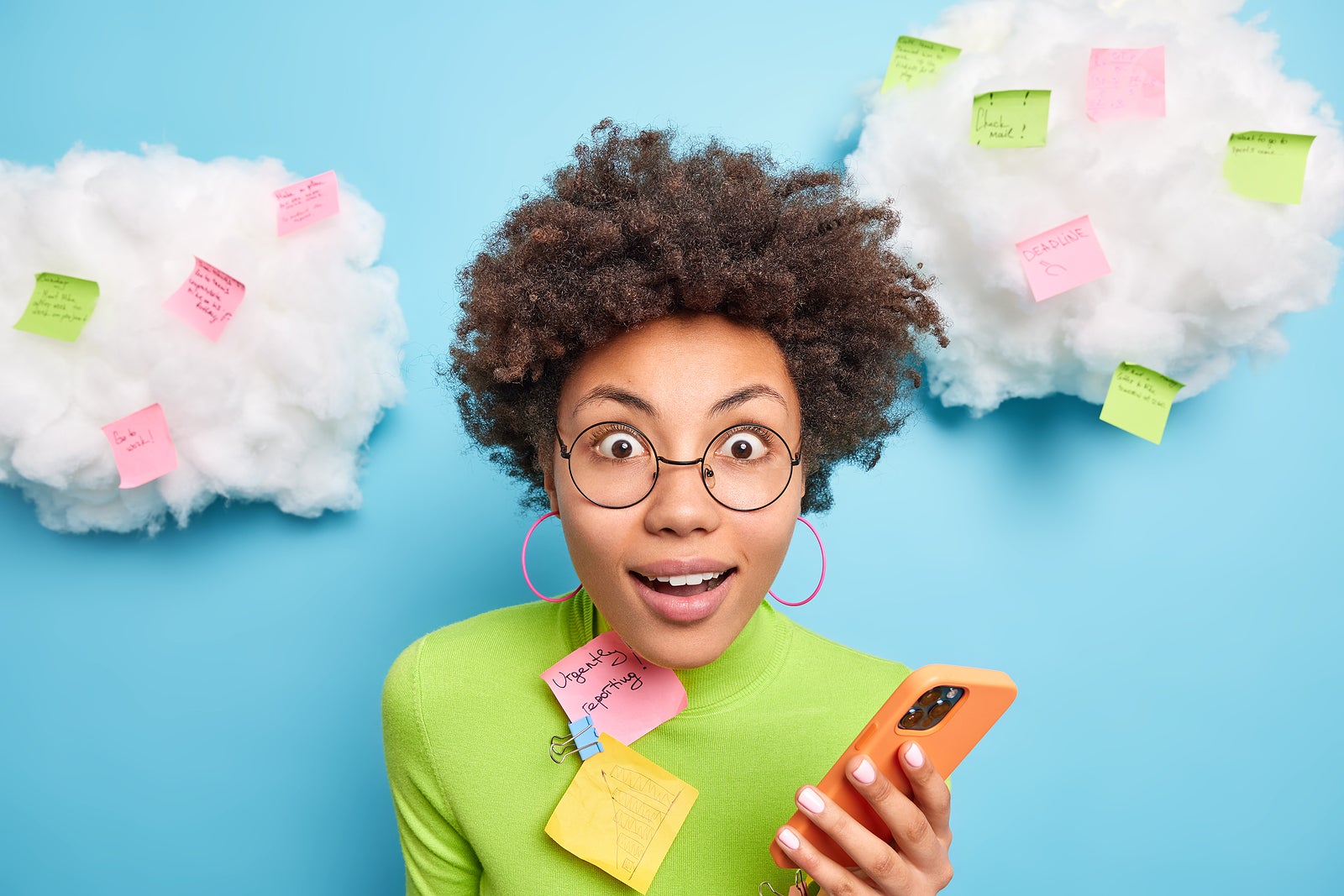 Close up shot of surprised curly woman stares surprisingly at camera holds mobile phone checks email box surrounded with memo notes makes list to do tries to be more productive poses indoor.