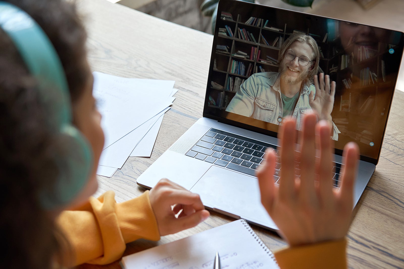 Hispanic teen girl school college student distance learning waving hand studying with online teacher on laptop screen. Elearning zoom video call, videoconference class with tutor. Over shoulder view.