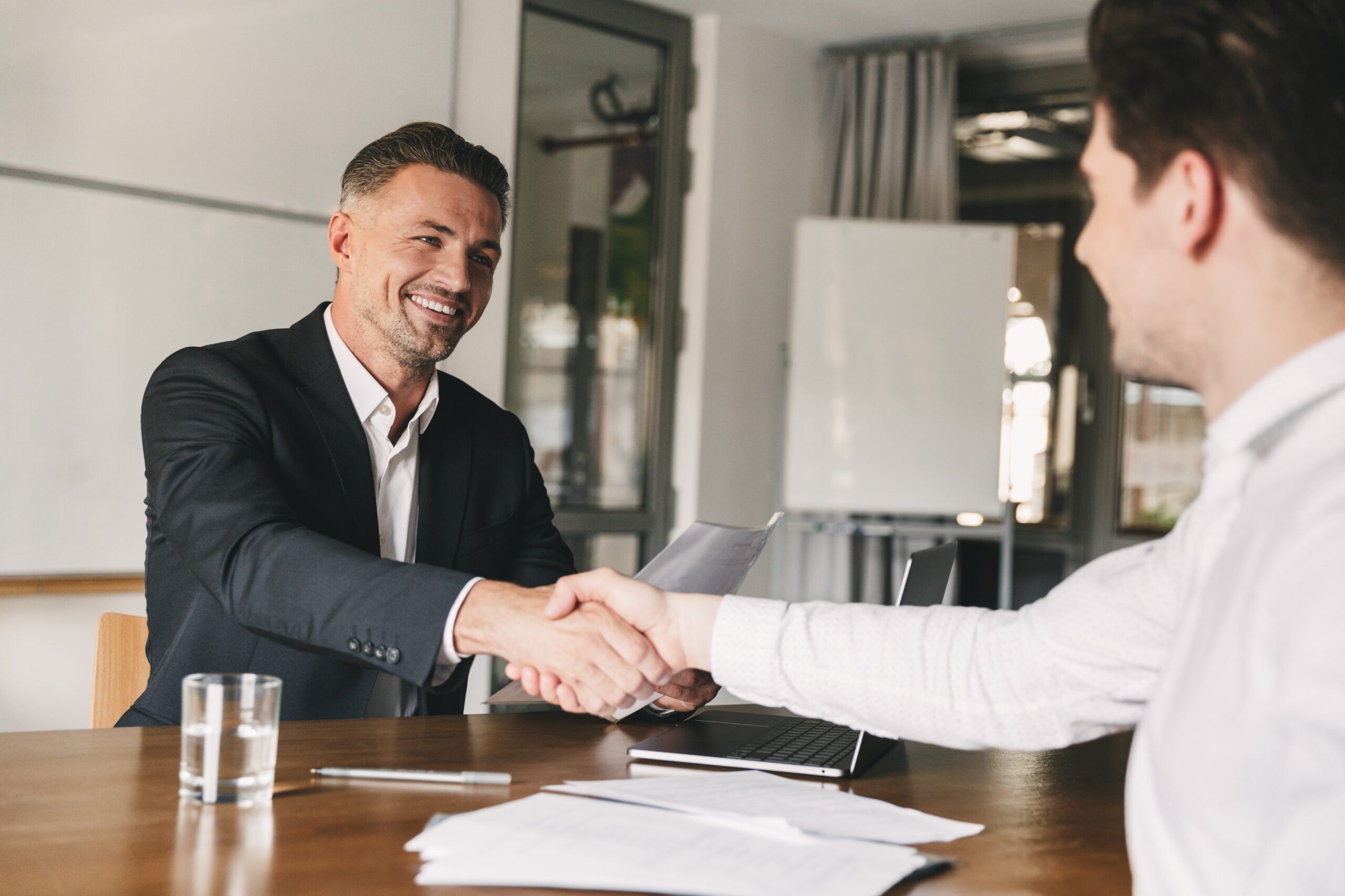 Business, career and placement concept - joyful handsome businessman 30s smiling and shaking hands with male candidate who was recruited during interview in office