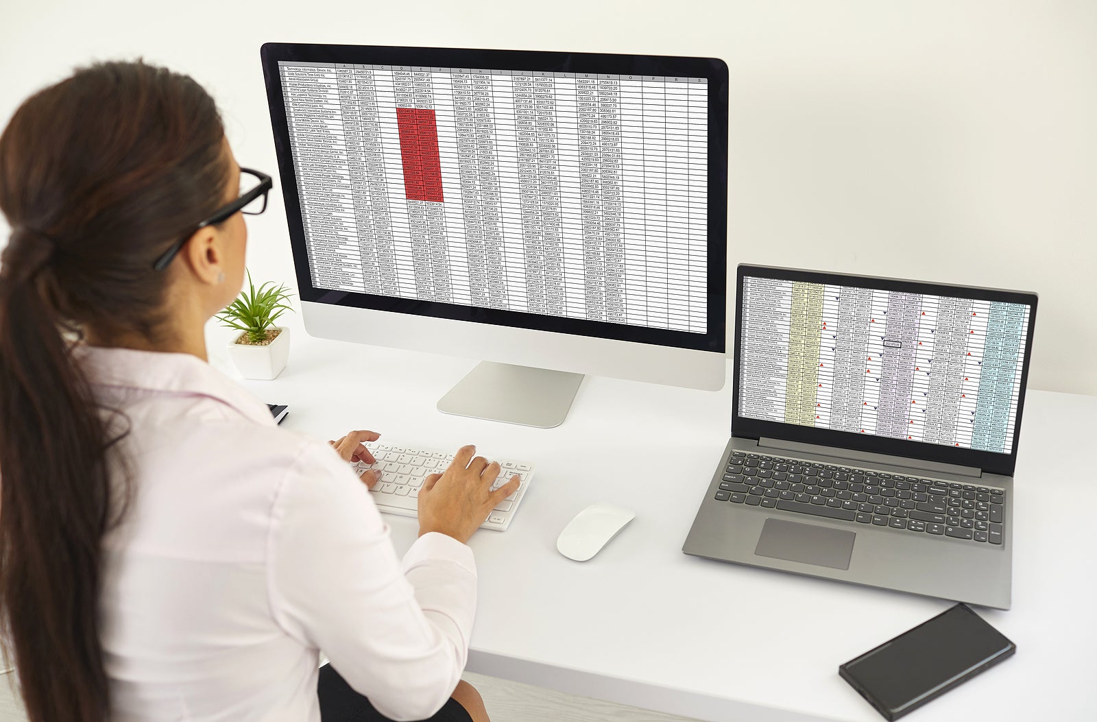 Woman sitting at office desk and working with data lists on desktop and laptop computers