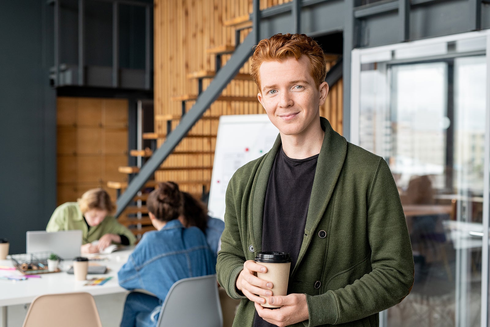 Young successful office manager in casualwear having drink in front of camera and looking at you against colleagues having meeting