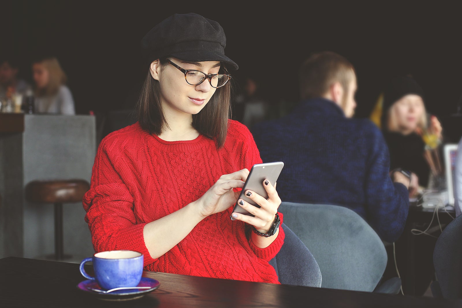 Young attractive millennial hipster brunette girl wearing baby blue shirt using tablet &amp; phone, sitting by big window of coffee shop / bar, drinking cappuccino. Copy space, front view.