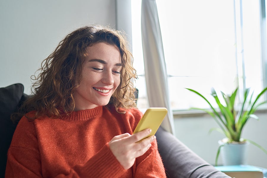 Smiling young woman sitting on couch using cell phone, happy lady holding smartphone, looking at cellphone doing online shopping in mobile apps ordering ecommerce products or checking social media.