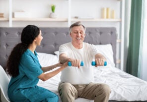 Young physiotherapist helping homebound elderly man to do physical exercises with dumbbells on bed at home