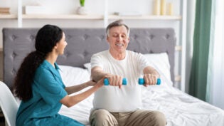 Young physiotherapist helping homebound elderly man to do physical exercises with dumbbells on bed at home