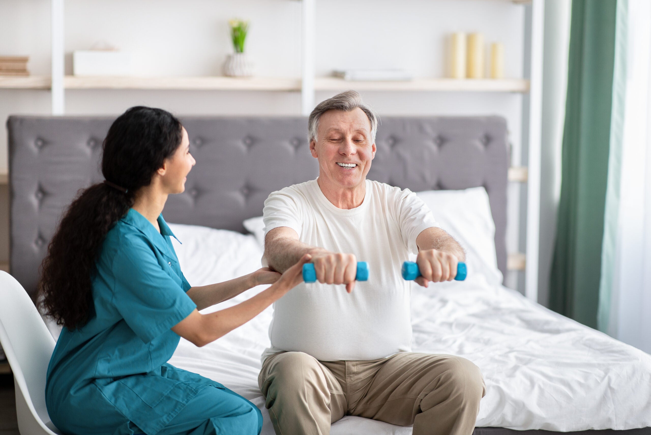 Young physiotherapist helping homebound elderly man to do physical exercises with dumbbells on bed at home Young physiotherapist helping homebound elderly man to do physical exercises with dumbbells on bed at home