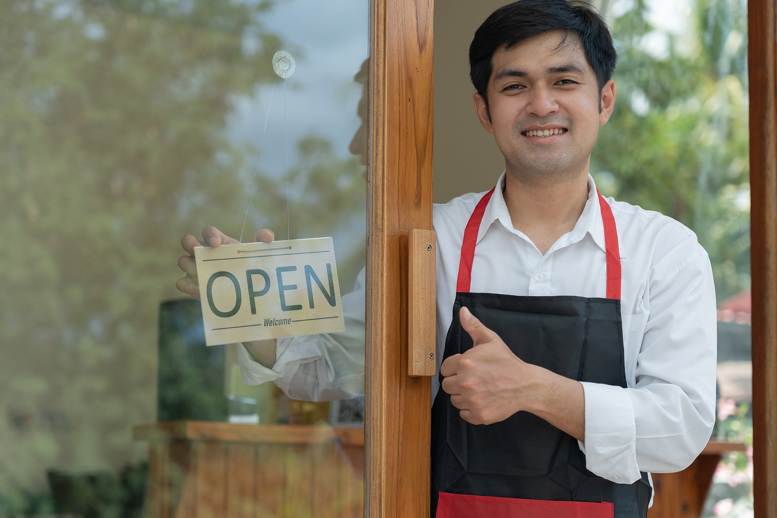 Business owner thumbs up to show his readiness to welcome customers. waitress man turning on open sign board on glass door in modern cafe coffee shop. New normal, small business, cafe, life style.