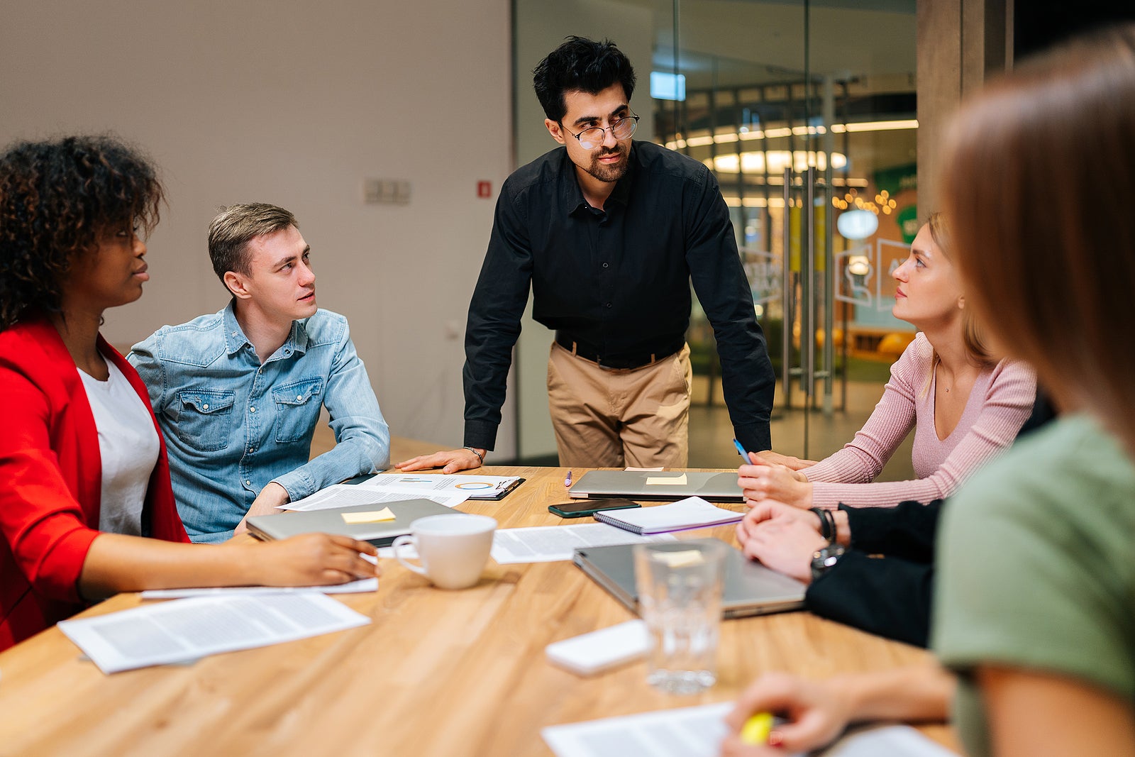 Portrait of confident business team leader standing surrounded by multiracial colleagues, confidently looking to staff. Multiethnic startup business team posing at table in modern meeting office room.