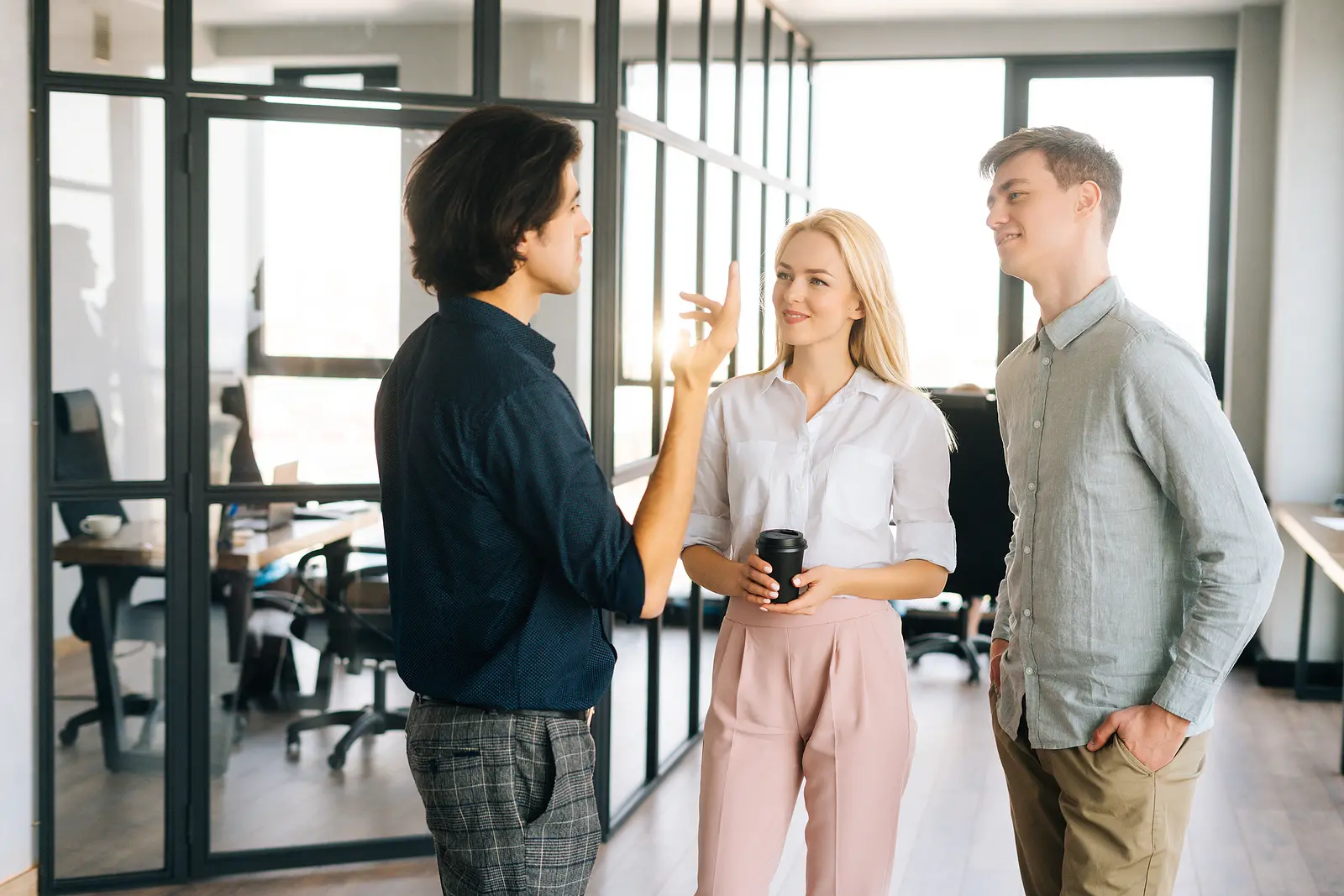 Portrait of three successful young colleagues standing in modern loft office talking and discussing business issues during coffee break. Male and female office staff standing and having conversation.