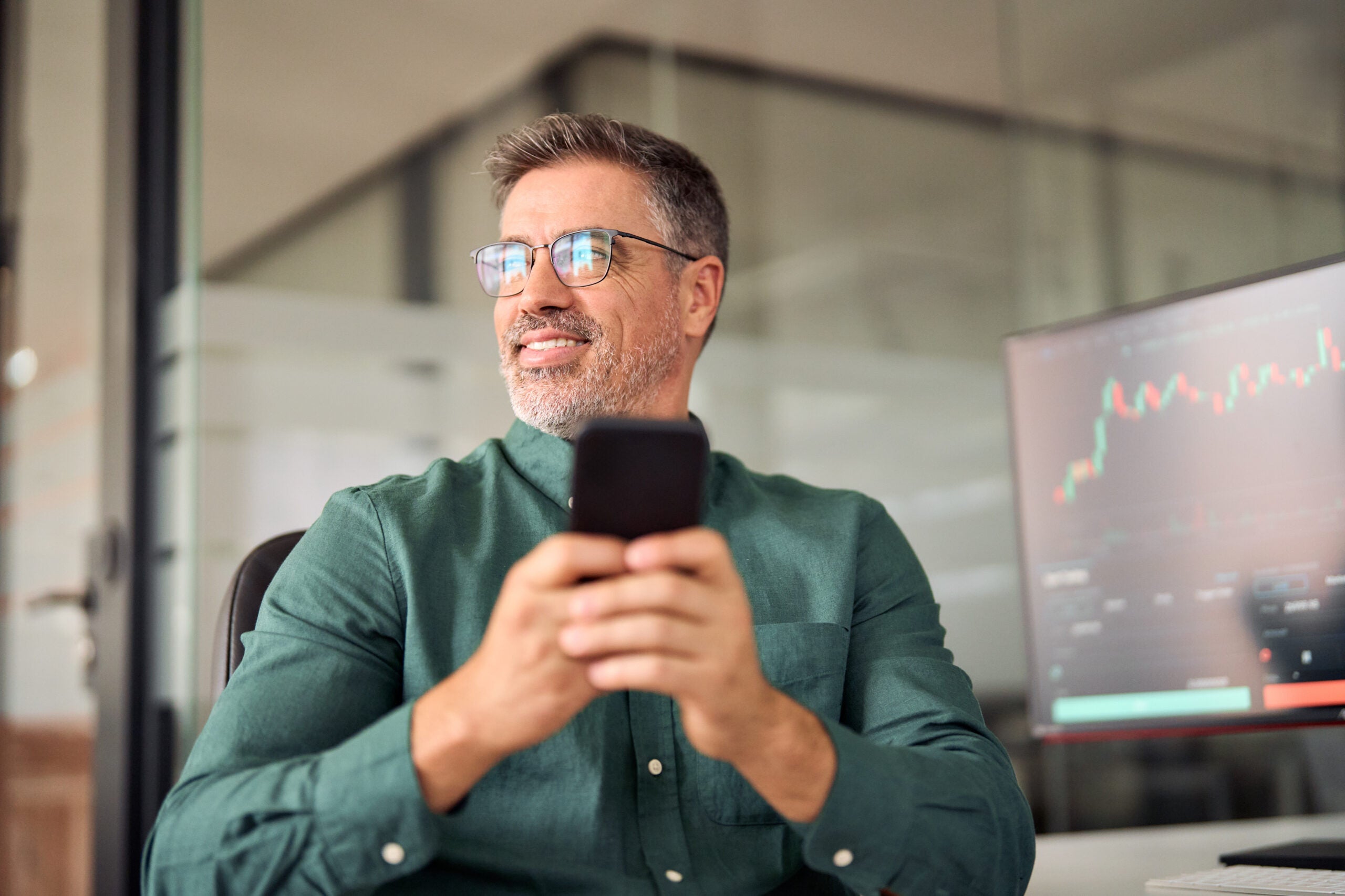 Crypto investor broker using smartphone trading app on cell phone. Middle aged stock trader analyzing financial trade stock market digital data on smartphone thinking of investing funds looking aside.