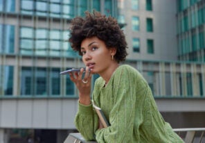 Horizontal shot of thoughtful curly haired woman uses cellphone for communication over speaker records voice message dressed casually poses against modern city building. Modern technologies.