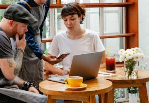 Three people collaborate around a laptop and smartphone at a small café table, with a cup of coffee in the foreground.
