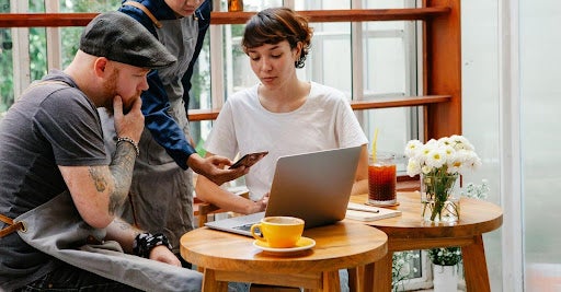 Three people collaborate around a laptop and smartphone at a small café table, with a cup of coffee in the foreground.
