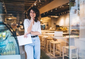Happy female manager with menu in hand standing in doorway of local cafeteria and smiling, pretty Caucaisan small owner enjoying business lifestyle for working with franchise takeaway industry