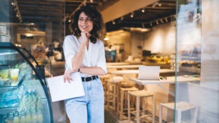 Happy female manager with menu in hand standing in doorway of local cafeteria and smiling, pretty Caucaisan small owner enjoying business lifestyle for working with franchise takeaway industry