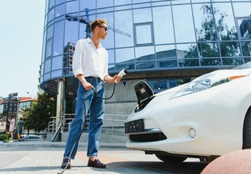 Man Holding Power Charging Cable For Electric Car In Outdoor Car Park. And he s going to connect the car to the charging station in the parking lot near the shopping center
