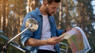 Focused man looking map of USA near scooter on asphalt road in blur forest. Concept of travelling, vacation, journey and trip. Young handsome caucasian guy. Sunny day