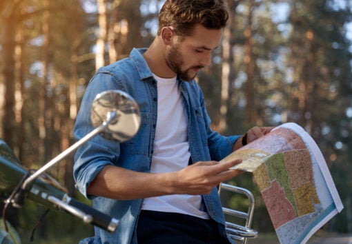 Focused man looking map of USA near scooter on asphalt road in blur forest. Concept of travelling, vacation, journey and trip. Young handsome caucasian guy. Sunny day