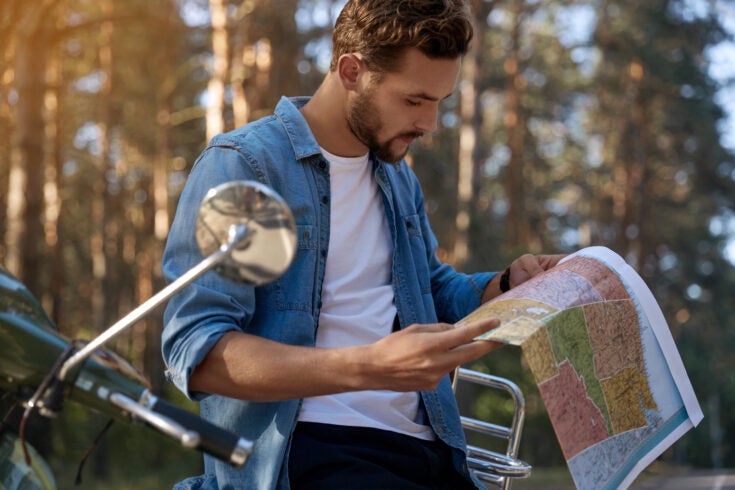 Focused man looking map of USA near scooter on asphalt road in blur forest. Concept of travelling, vacation, journey and trip. Young handsome caucasian guy. Sunny day