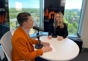 Man in orange jacket interviewing blonde woman with glasses